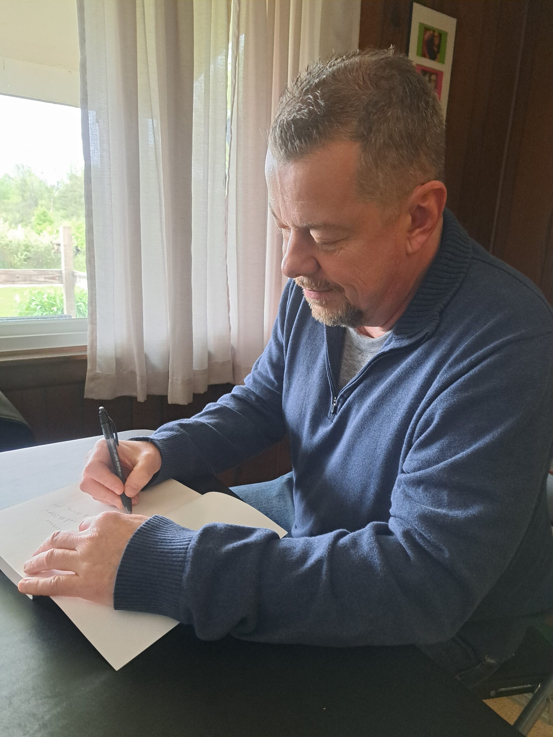 Author Mark Saluke signing a copy of Lucy’s Way at a table by a window.