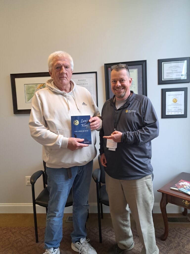Former counselor standing next to author Mark Saluke while holding his book Lucy’s Way