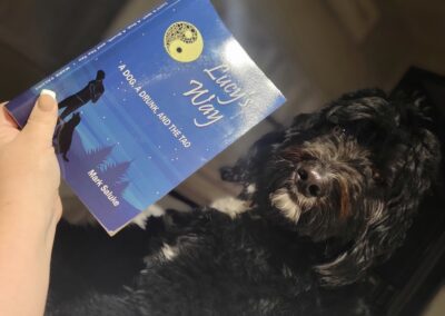 Person holding a copy of Lucy’s Way by Mark Saluke next to their dog
