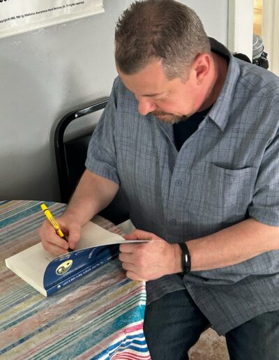 Author Mark Saluke signing a copy of Lucy’s Way at a table.